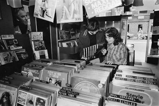 Two people are standing in a record store, flipping through vinyl records. Various music genres and album covers are visible, organized in rows. The space has a nostalgic, retro feel, with dim lighting and posters on the walls.