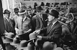 A group of men, all wearing suits and hats, are gathered in a factory-like setting, examining hats. The room is filled with shelves of hats in various stages of completion. The attire and setting suggest a mid-20th-century time period, possibly in the manufacturing or quality control section of a hat factory.