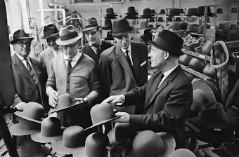 A group of men, all wearing suits and hats, are gathered in a factory-like setting, examining hats. The room is filled with shelves of hats in various stages of completion. The attire and setting suggest a mid-20th-century time period, possibly in the manufacturing or quality control section of a hat factory.