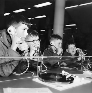 Four boys are sitting at a table with headphones, listening intently. The setup includes a record player, indicating they are likely listening to music or an audio recording. The environment suggests a public exhibit or a learning station.