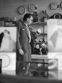 A man in a suit is examining a clock in a shop filled with various clocks displayed on shelves and walls. The setting likely captures a timepiece store with a selection of analog clocks. The background includes plants and signage, suggesting a well-organized retail space.