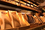 A display of elegantly packaged golden chocolate bars prominently arranged, with a shelf lined with more products in the background. The setting suggests a specialty chocolate shop or a gourmet store with a warm, inviting atmosphere.