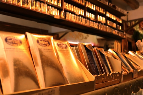 A display of elegantly packaged golden chocolate bars prominently arranged, with a shelf lined with more products in the background. The setting suggests a specialty chocolate shop or a gourmet store with a warm, inviting atmosphere.