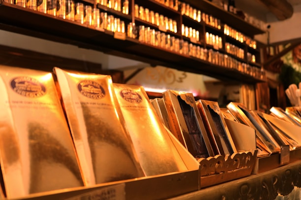 A display of elegantly packaged golden chocolate bars prominently arranged, with a shelf lined with more products in the background. The setting suggests a specialty chocolate shop or a gourmet store with a warm, inviting atmosphere.