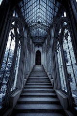 An intricately designed, narrow staircase ascends towards a large, ornate wooden door. The surrounding architecture is Gothic, with tall, arched windows lining the sides, allowing soft light to filter through. The ceiling features elaborate latticework in a vaulted design.