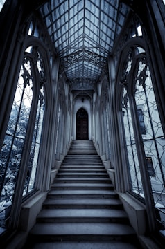 An intricately designed, narrow staircase ascends towards a large, ornate wooden door. The surrounding architecture is Gothic, with tall, arched windows lining the sides, allowing soft light to filter through. The ceiling features elaborate latticework in a vaulted design.