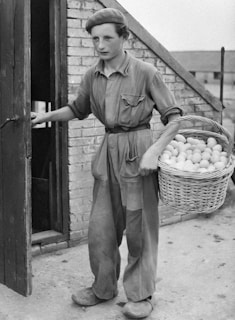 A young person is dressed in loose-fitting work attire, holding a wicker basket filled with eggs. They are standing outside a building with a brick wall and an open wooden door, suggesting a farm or rural setting.