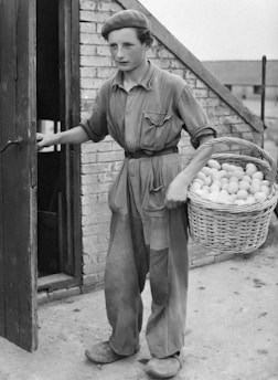 A young person is dressed in loose-fitting work attire, holding a wicker basket filled with eggs. They are standing outside a building with a brick wall and an open wooden door, suggesting a farm or rural setting.