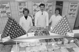 Three people in white uniforms, including two women and one man, are standing behind a meat counter filled with various meat products and sausages. The counter features neatly arranged piles of sausages and what appears to be bread or buns. There are several price tags displayed on the counter. Behind them, there are signs with prices for different products on the tiled wall.