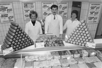 Three people in white uniforms, including two women and one man, are standing behind a meat counter filled with various meat products and sausages. The counter features neatly arranged piles of sausages and what appears to be bread or buns. There are several price tags displayed on the counter. Behind them, there are signs with prices for different products on the tiled wall.
