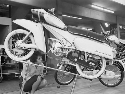 A vintage motorcycle displayed on a stand with a woman crouching next to it, observing the details. The motorcycle has a classic design with sleek curves and chrome accents, and is situated indoors, likely in a showroom or exhibition space.