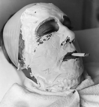 A person is lying down with a facial mask applied, mostly covering the face except for the eyes and mouth. The person is smoking a cigarette, which protrudes from the mask. A headband holds back hair, and a towel is wrapped around the neck.