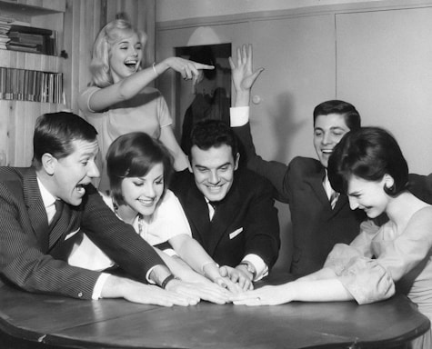 A group of six people are gathered around a table, appearing to engage in a social game or activity. They are smiling and laughing, showing a sense of enjoyment and camaraderie. The setting seems to be indoors with books on a shelf in the background.