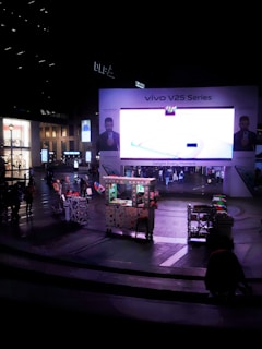 A large electronic billboard displays an advertisement for the Vivo V25 Series in a bustling urban plaza. People are walking around, and a small cart is visible in the foreground, adorned with lights and decorations. Surrounding buildings are softly illuminated, contributing to the nighttime atmosphere.