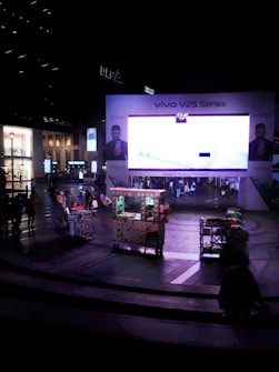 A large electronic billboard displays an advertisement for the Vivo V25 Series in a bustling urban plaza. People are walking around, and a small cart is visible in the foreground, adorned with lights and decorations. Surrounding buildings are softly illuminated, contributing to the nighttime atmosphere.