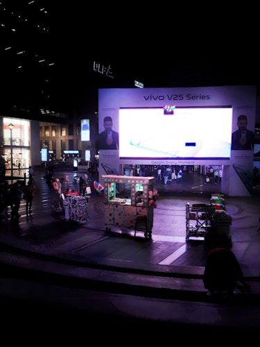 A large electronic billboard displays an advertisement for the Vivo V25 Series in a bustling urban plaza. People are walking around, and a small cart is visible in the foreground, adorned with lights and decorations. Surrounding buildings are softly illuminated, contributing to the nighttime atmosphere.