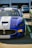 A blue Maserati race car is parked in a lot, featuring prominent brand decals like Pirelli on the front bumper. The car has an aggressive design with a large grille, aerodynamic elements, and racing stripes.