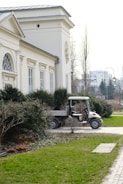 A small utility vehicle is parked beside a neoclassical building with arched windows and ornate design. The foreground features neatly trimmed bushes and a manicured lawn, while the background shows a quiet urban street lined with trees and lampposts.