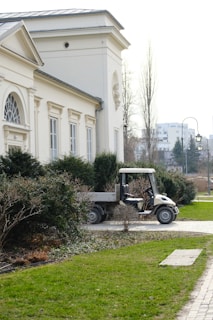 A small utility vehicle is parked beside a neoclassical building with arched windows and ornate design. The foreground features neatly trimmed bushes and a manicured lawn, while the background shows a quiet urban street lined with trees and lampposts.