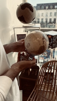 A person is holding a small globe while standing near a window. Another globe sits on a stand nearby. The setting appears to be indoors with natural light streaming in from outside. A wicker chair and some boxes are visible in the background.