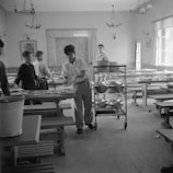 A group of young people are in a dining hall with long tables and benches. One person is handling plates and utensils, possibly clearing the table. A trolley with stacked plates is nearby, suggesting a communal dining setting.