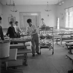 A group of young people are in a dining hall with long tables and benches. One person is handling plates and utensils, possibly clearing the table. A trolley with stacked plates is nearby, suggesting a communal dining setting.