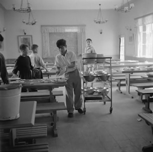 A group of young people are in a dining hall with long tables and benches. One person is handling plates and utensils, possibly clearing the table. A trolley with stacked plates is nearby, suggesting a communal dining setting.