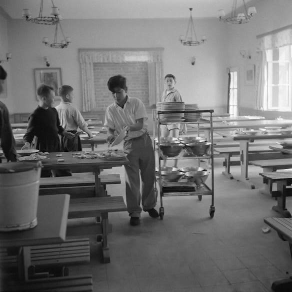A group of young people are in a dining hall with long tables and benches. One person is handling plates and utensils, possibly clearing the table. A trolley with stacked plates is nearby, suggesting a communal dining setting.