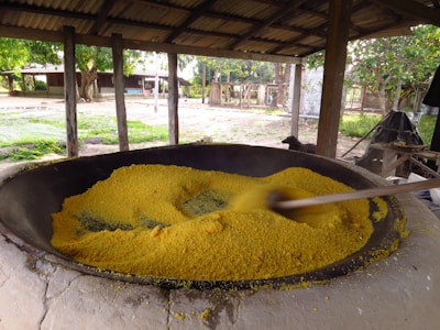 A large outdoor traditional cooking setup with a wide black pan filled with bright yellow grains, being stirred by a large wooden stick. There is a rustic, open-sided shelter surrounding the pan, with wooden beams and a thatched roof. In the background, there are trees, greenery, and some structures that suggest a rural setting.