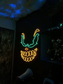 A dark room is illuminated by a neon light display depicting a menacing, glowing face with exaggerated eyes and a broad, toothy grin. The neon colors are green and yellow, creating an eerie atmosphere. On the ceiling, there are cloud-like structures with a hint of blue light enhancing the ambience. The room contains several metallic objects and toys, including a megaphone and small model cars. The wall features a design resembling a cityscape.