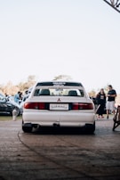 A white Mitsubishi car is parked on a paved area with a license plate that reads 'Schwanz'. In the background, several people and cars are visible, indicating a possible car gathering or event. The sky is clear, and it seems to be daytime.