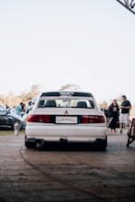 A white Mitsubishi car is parked on a paved area with a license plate that reads 'Schwanz'. In the background, several people and cars are visible, indicating a possible car gathering or event. The sky is clear, and it seems to be daytime.