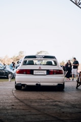 A white Mitsubishi car is parked on a paved area with a license plate that reads 'Schwanz'. In the background, several people and cars are visible, indicating a possible car gathering or event. The sky is clear, and it seems to be daytime.