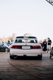 A white Mitsubishi car is parked on a paved area with a license plate that reads 'Schwanz'. In the background, several people and cars are visible, indicating a possible car gathering or event. The sky is clear, and it seems to be daytime.
