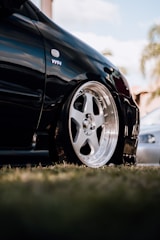 A close-up view of a sleek black car featuring a shiny, polished alloy wheel. The surroundings include blurred elements suggesting a grassy area and some trees in the distance. The polished surface of the car reflects its high-quality finish.