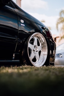A close-up view of a sleek black car featuring a shiny, polished alloy wheel. The surroundings include blurred elements suggesting a grassy area and some trees in the distance. The polished surface of the car reflects its high-quality finish.