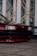 A close-up of the front section of a red car with sleek and glossy paint. The car is parked on a tiled surface with a backdrop of modern architectural elements featuring large glass windows and metal frames.