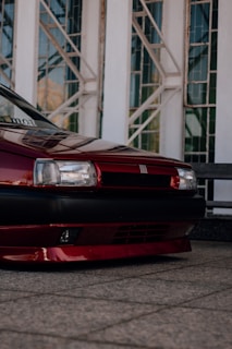 A close-up of the front section of a red car with sleek and glossy paint. The car is parked on a tiled surface with a backdrop of modern architectural elements featuring large glass windows and metal frames.