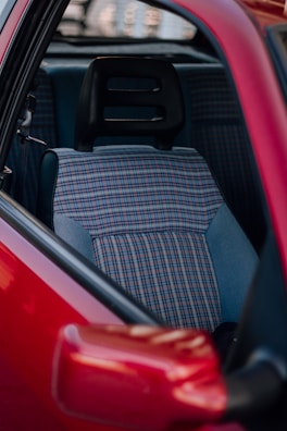 A close-up view of a car's interior, featuring a seat with a plaid fabric pattern. The seat includes a black headrest and is surrounded by a red car door.