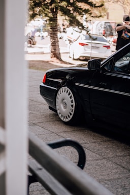 A black Volkswagen car with distinctive round wheel covers is parked on a paved area. In the background, there are several other vehicles, including a white car, and a person standing near them. There is a tree in the scene and visible shadows indicating a sunny day.