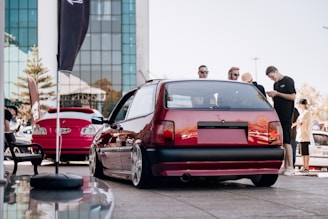 A group of people stand near a red hatchback car with modified wheels parked on a street. Another red car is parked nearby, and a modern glass building is in the background. The weather appears sunny.