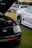 A black Toyota car with its hood open is parked on the grass. A white sports car is visible alongside, slightly out of focus, showing a sleek design and shiny body. A third car is in the background, partially visible. The scene is an outdoor setting with a relaxed atmosphere.