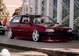 A red, vintage-style hatchback car with white wheels is parked on a concrete platform. The background features a building with large windows and architectural elements.