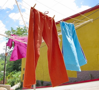 Colorful clothes hang on a clothesline under a bright sky. An orange pair of pants and a blue garment are prominently featured, along with a pink shirt. The background includes a vivid yellow wall and a view of greenery and a street.