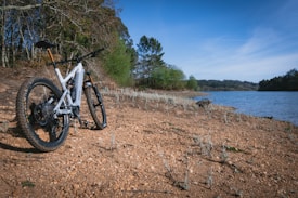 A mountain bike is positioned on a sandy path near a serene body of water. Trees and shrubs can be seen to the left, with a clear blue sky above, adding to the tranquil atmosphere.