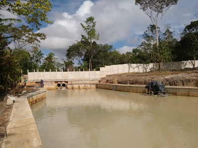 A small body of water surrounded by concrete walls, with construction pipes visible at the far end. Trees and dense vegetation are spread along the background, with a partly cloudy sky above. Construction materials and equipment are scattered around the site.