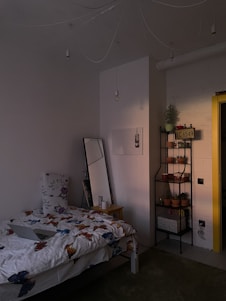A cozy bedroom with dim lighting, featuring a bed with butterfly-patterned bedding and a large floral pillow. Next to the bed is a wooden nightstand with a mug and a tall mirror leaning against the wall. A black metal shelf holds multiple potted plants and a decorative license plate. The wall is painted white, and the yellow doorframe adds a pop of color.
