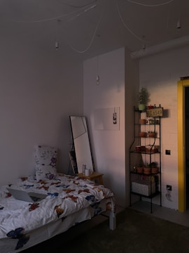 A cozy bedroom with dim lighting, featuring a bed with butterfly-patterned bedding and a large floral pillow. Next to the bed is a wooden nightstand with a mug and a tall mirror leaning against the wall. A black metal shelf holds multiple potted plants and a decorative license plate. The wall is painted white, and the yellow doorframe adds a pop of color.