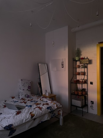 A cozy bedroom with dim lighting, featuring a bed with butterfly-patterned bedding and a large floral pillow. Next to the bed is a wooden nightstand with a mug and a tall mirror leaning against the wall. A black metal shelf holds multiple potted plants and a decorative license plate. The wall is painted white, and the yellow doorframe adds a pop of color.