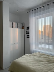 A minimalist bedroom features a cozy bed with a cream-colored fluffy blanket. To the side, there is a large white wardrobe with storage boxes on top. Sheer white curtains cover a window, allowing soft natural light to filter in. The room's color palette is neutral, and the view outside shows buildings reflecting the warm glow of sunset.